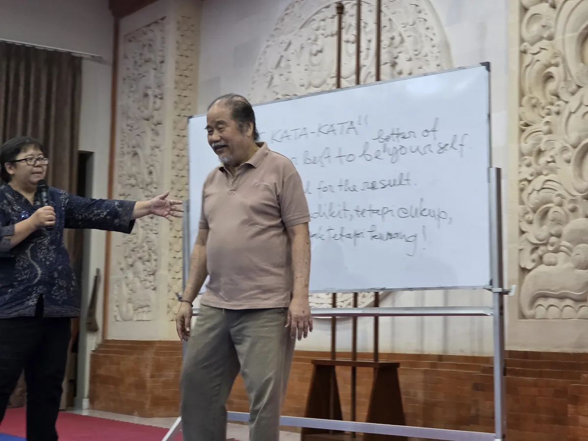 A host addressing the group beside a whiteboard written in a mixture of Indonesian and English